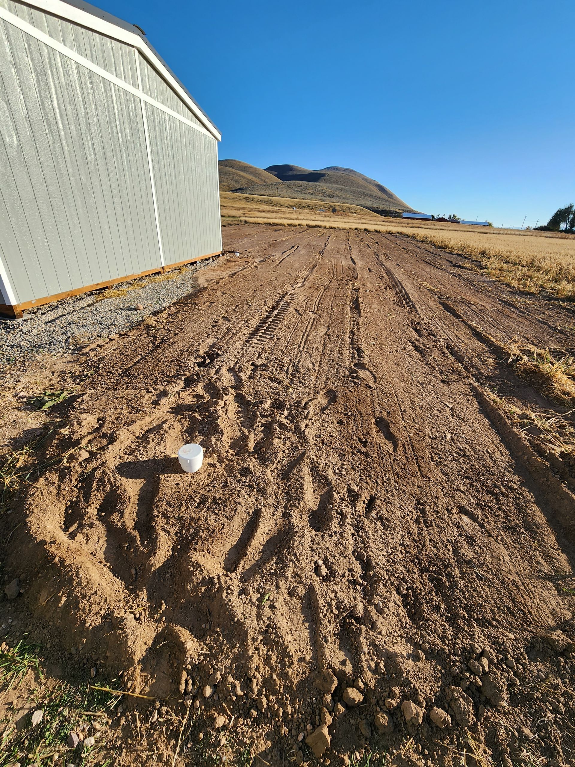 Rutted dirt path beside a white building under a clear blue sky, with hills in the distance