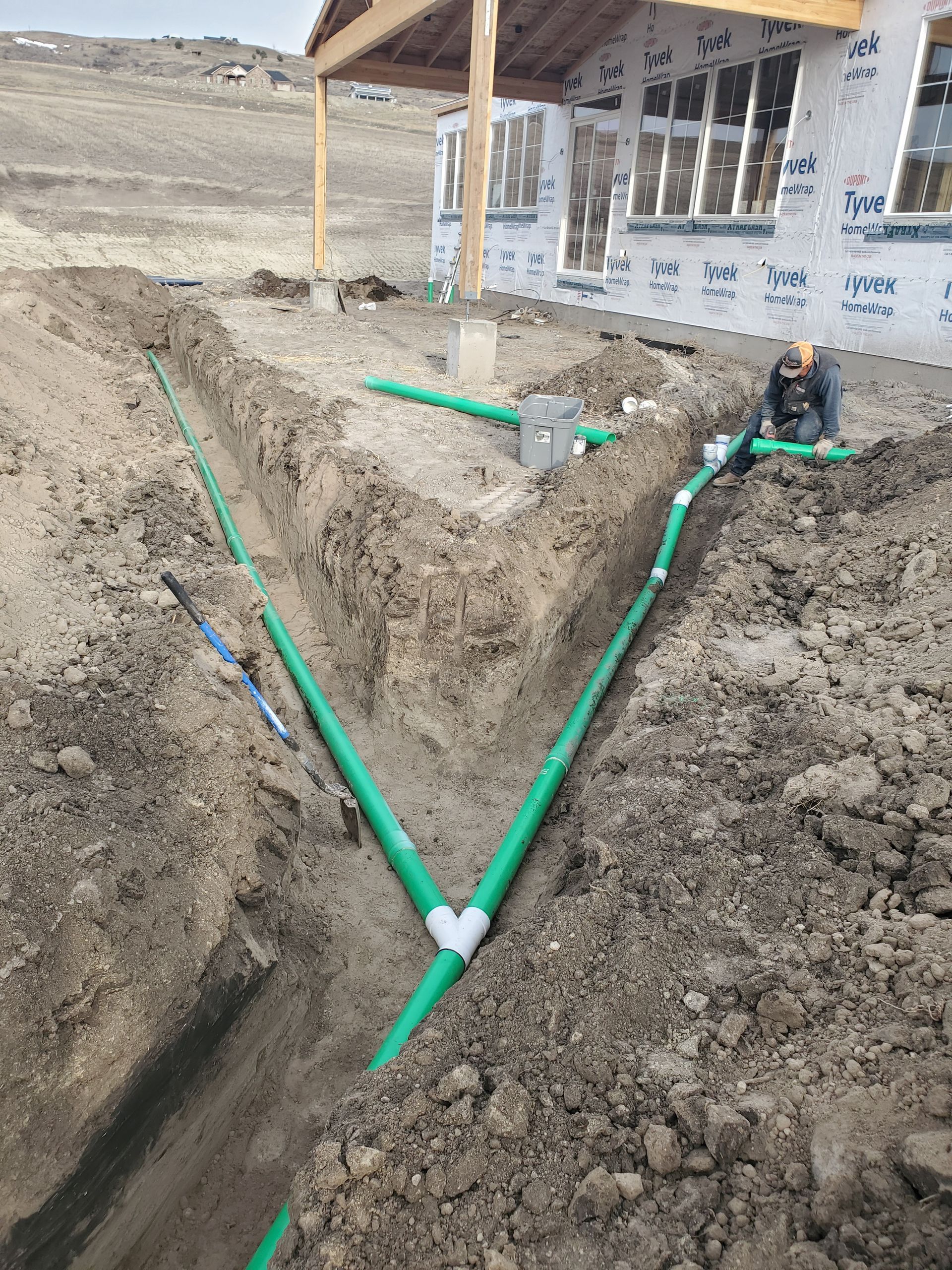 Green pipes laid in a V-shaped trench beside a house under construction.