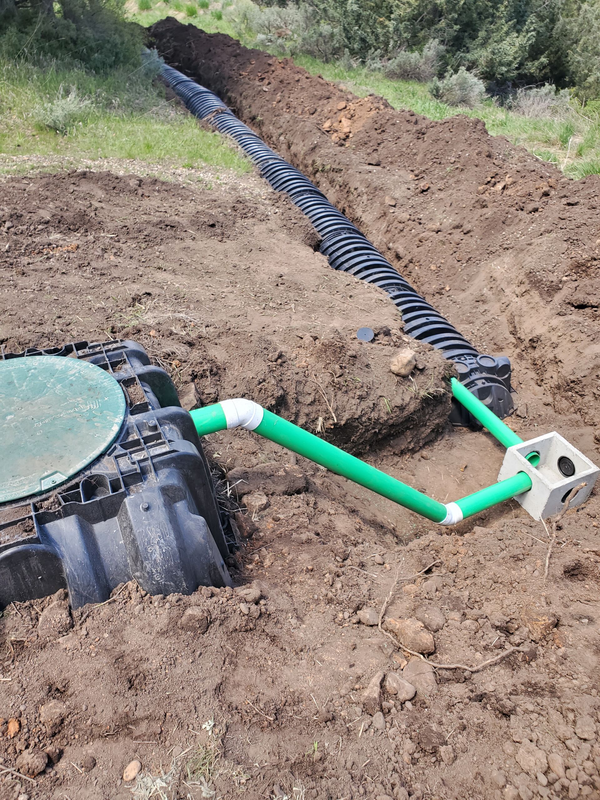 Septic tank installation trench with black corrugated pipe and green PVC pipe in a dirt yard
