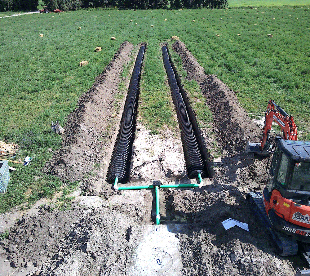 Trench excavation in a grassy field, with a red backhoe and black drainage pipes laid in parallel.