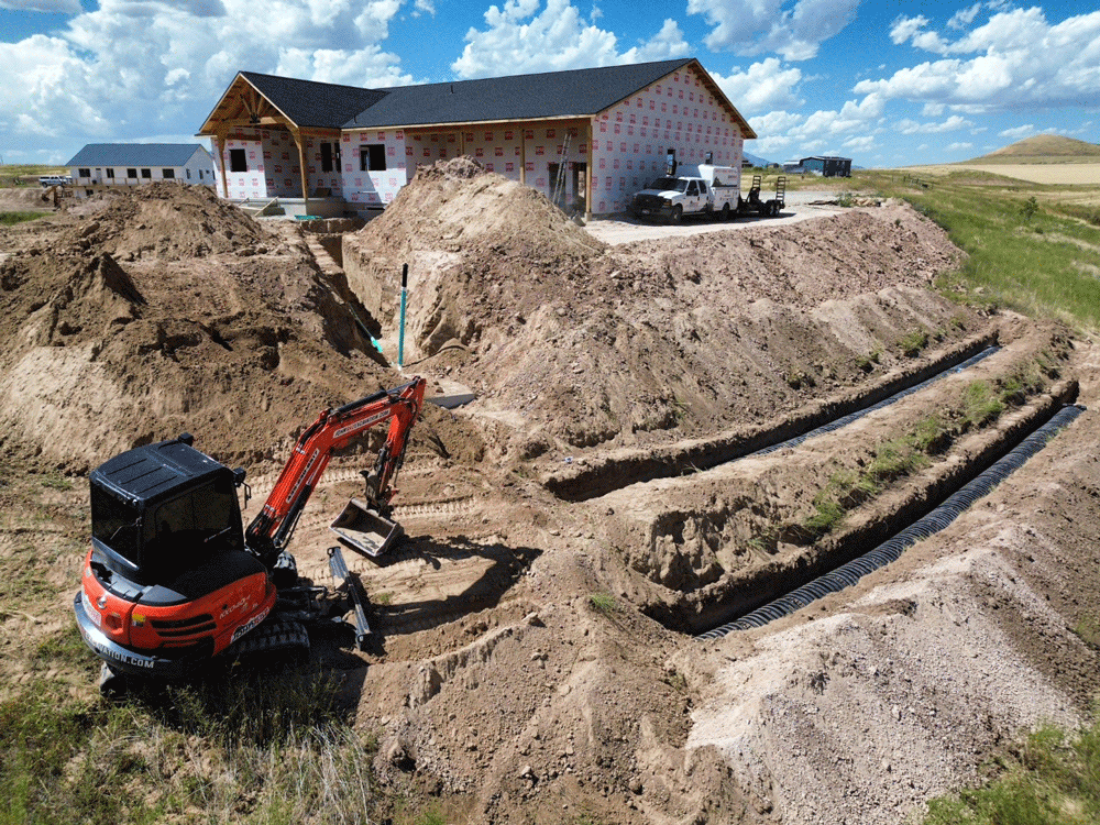 Excavator digging a dirt trench near a partially built house on a sandy construction site under a blue sky