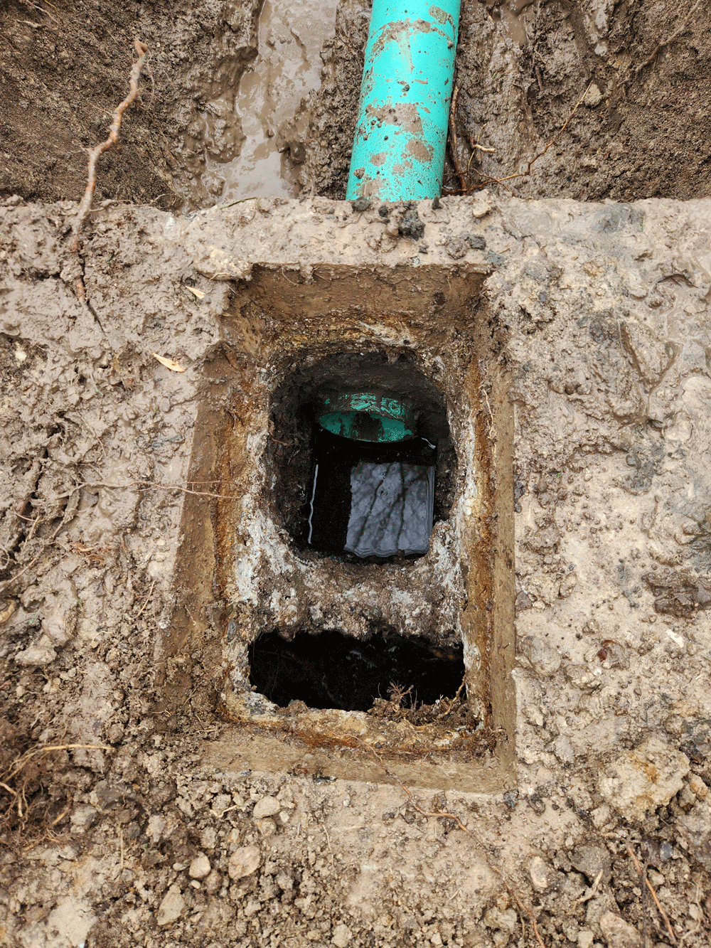 Rectangular concrete utility opening in dirt with a green pipe and dark interior underneath