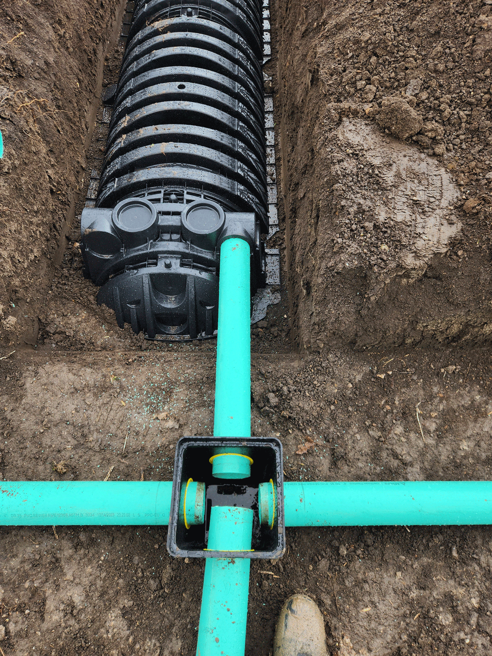 Green PVC pipes connected to a black corrugated drain pipe in a dirt trench.