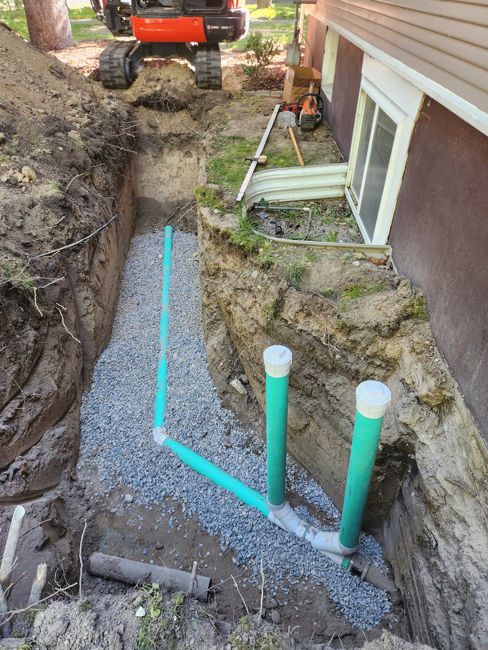 Backyard trench with gravel, green pipes, and an excavator in the distance next to a house.