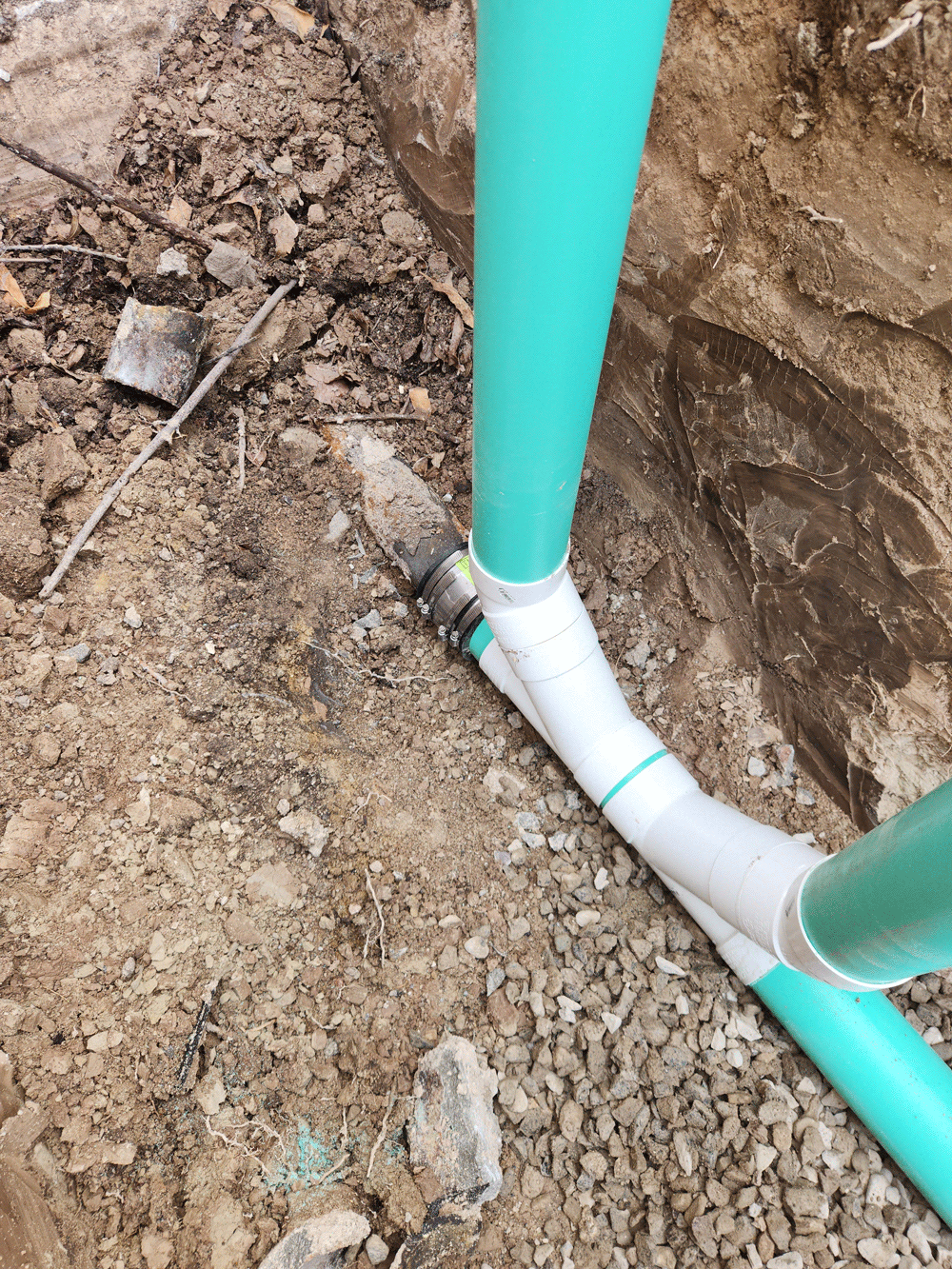 Turquoise pipes and white PVC drainage line in an excavated dirt trench