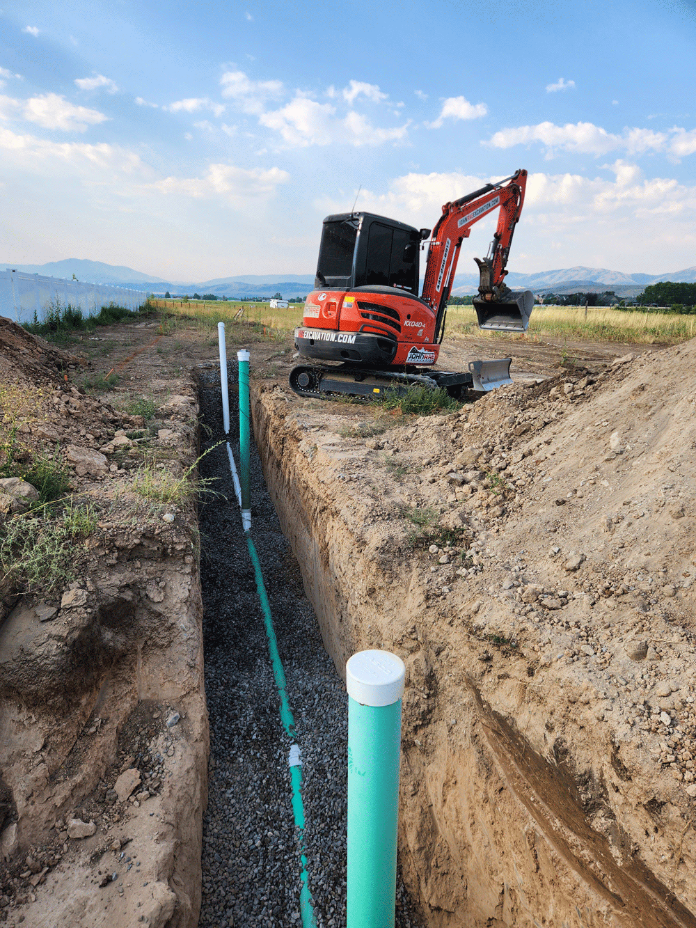 Orange excavator digging a trench with green pipes in a rural construction site