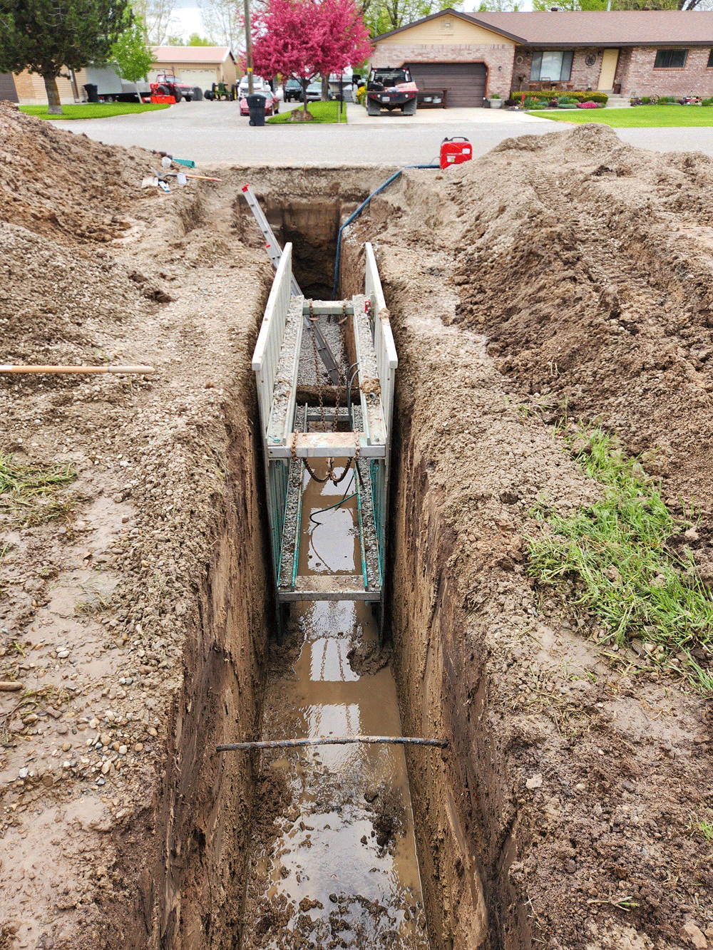 Excavated trench exposing a utility box and pipes in a muddy residential yard.