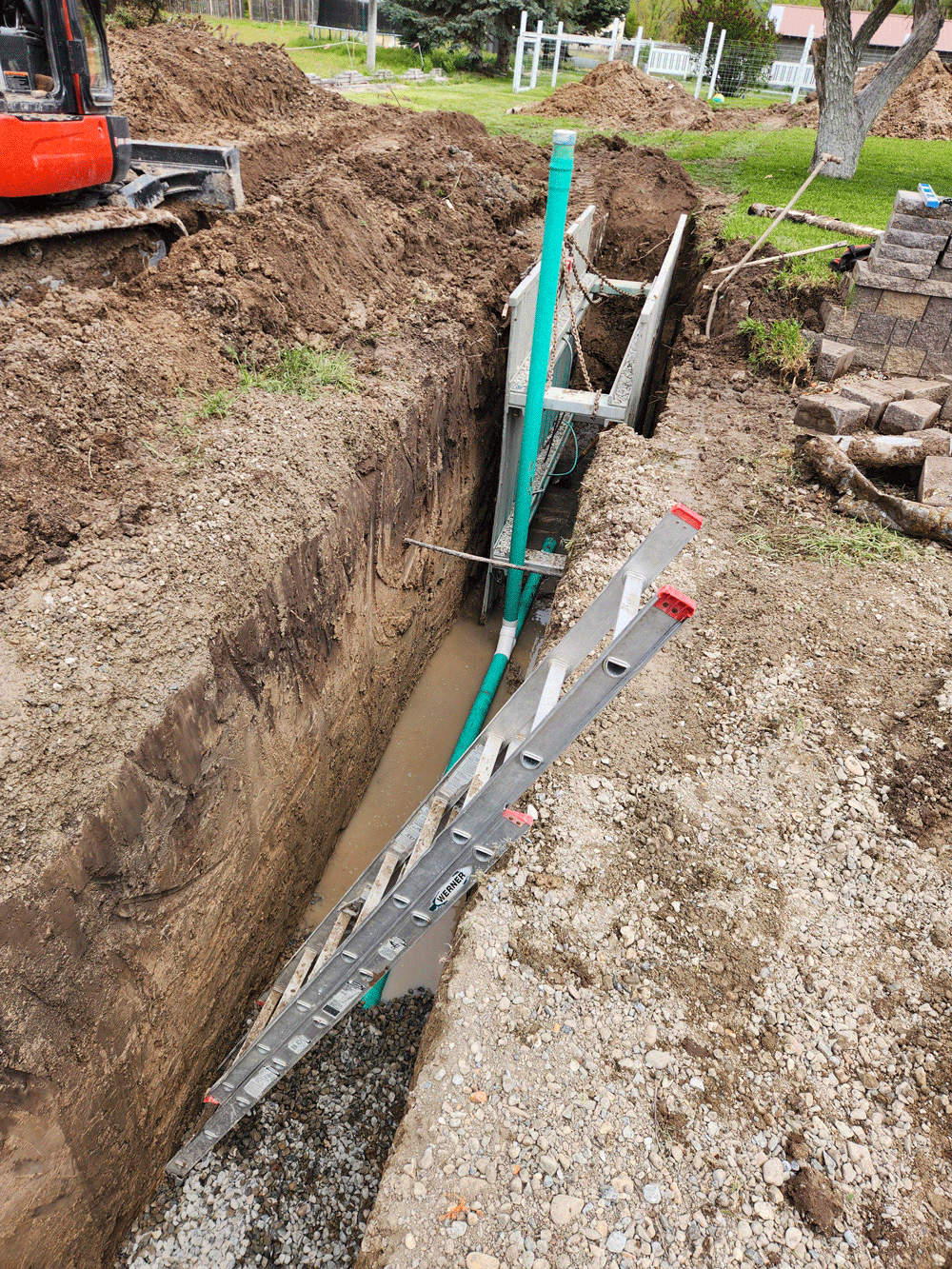 Excavator digging a trench beside a green post and gray drainage pipe in a gravel yard