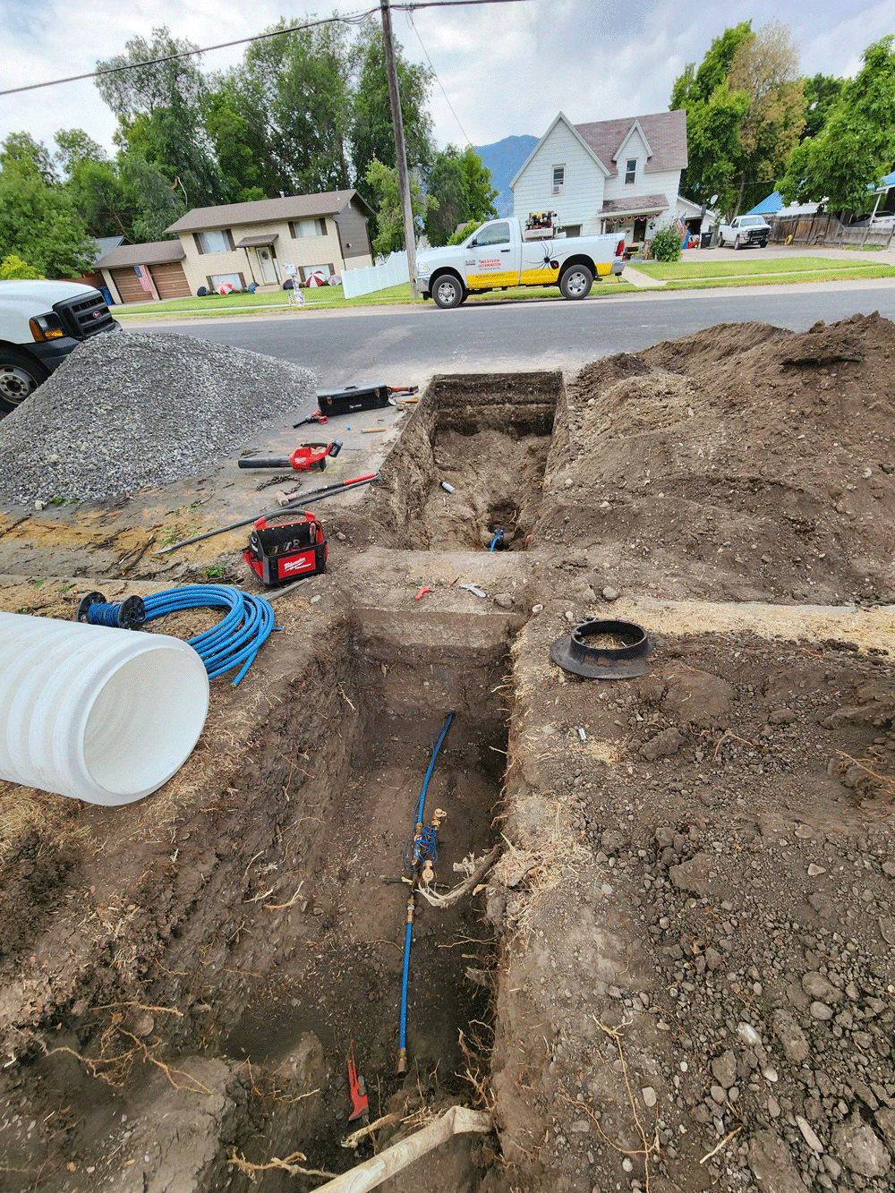 Road excavation trench with pipes and tools beside a suburban street, near parked vehicles and houses