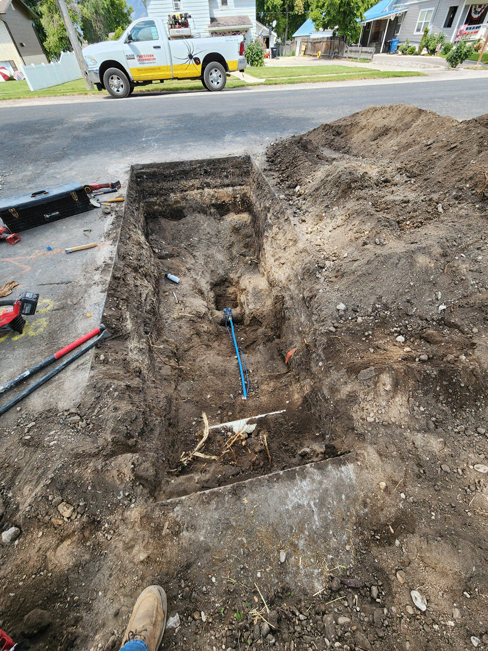 Open trench in a driveway with exposed pipes and dirt piles beside a parked pickup truck.