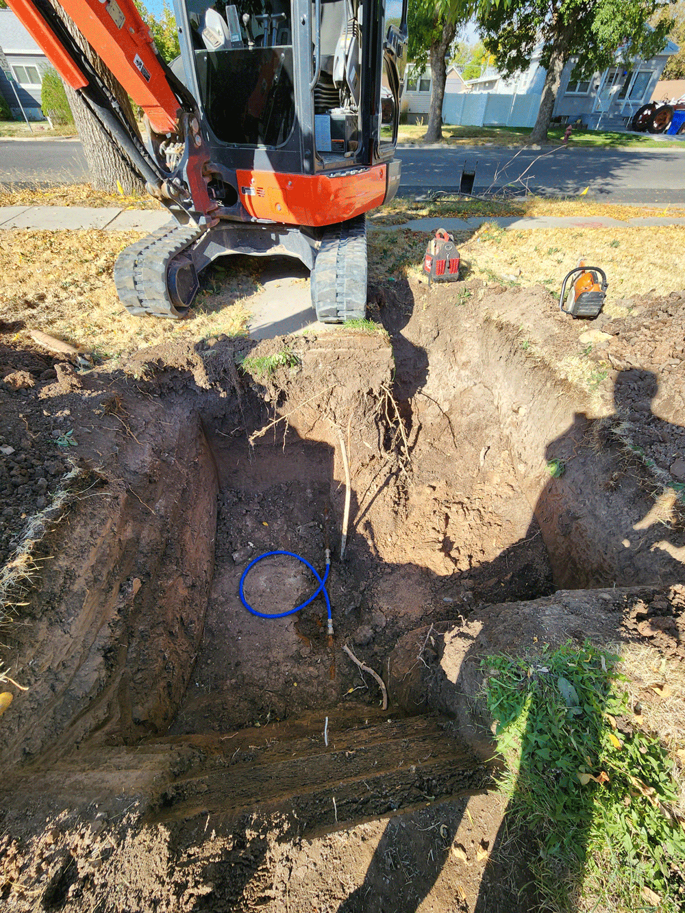 Excavator digging a deep trench in a yard with exposed dirt and utility tools nearby