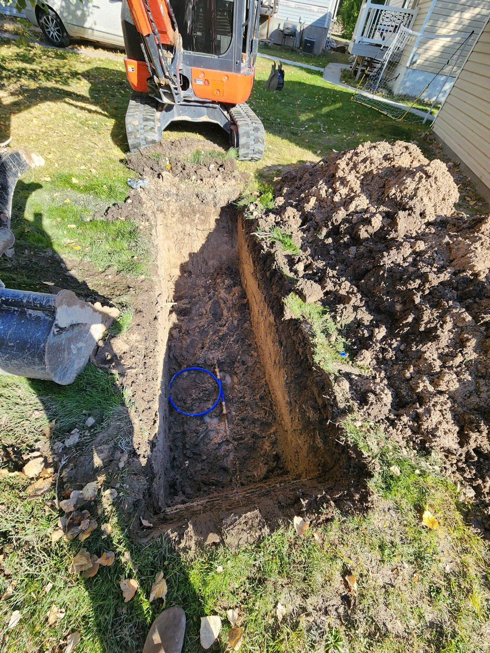 A trench being dug in a grassy yard with an orange excavator and piles of dirt beside it.