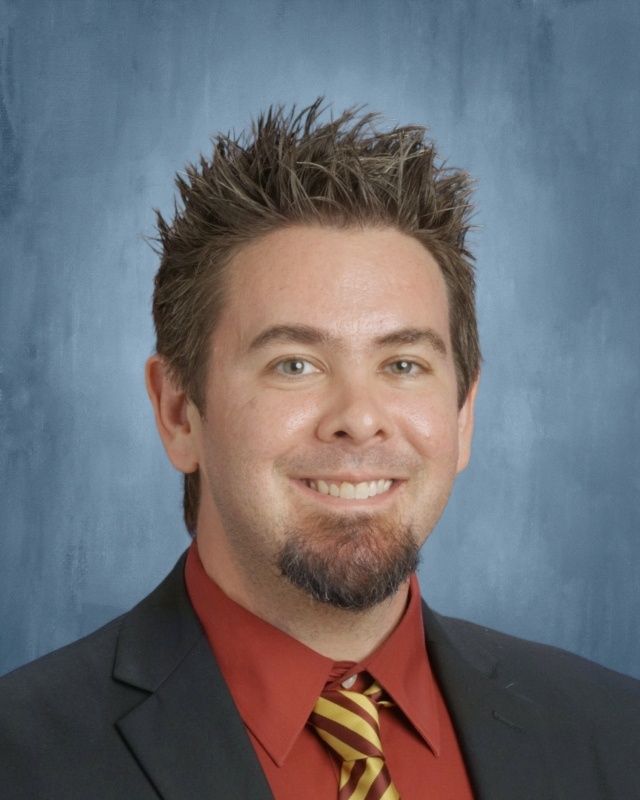 Man with spiky brown hair, goatee, smiling, wearing red shirt, tie, and navy blazer.