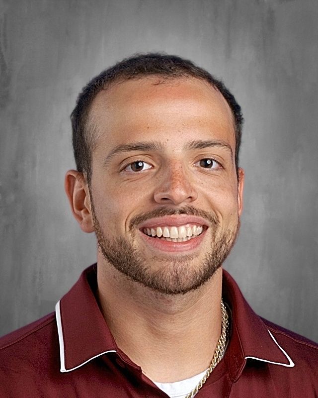 A young man with curly brown hair and a beard, wearing a pink polo, smiles at the camera.
