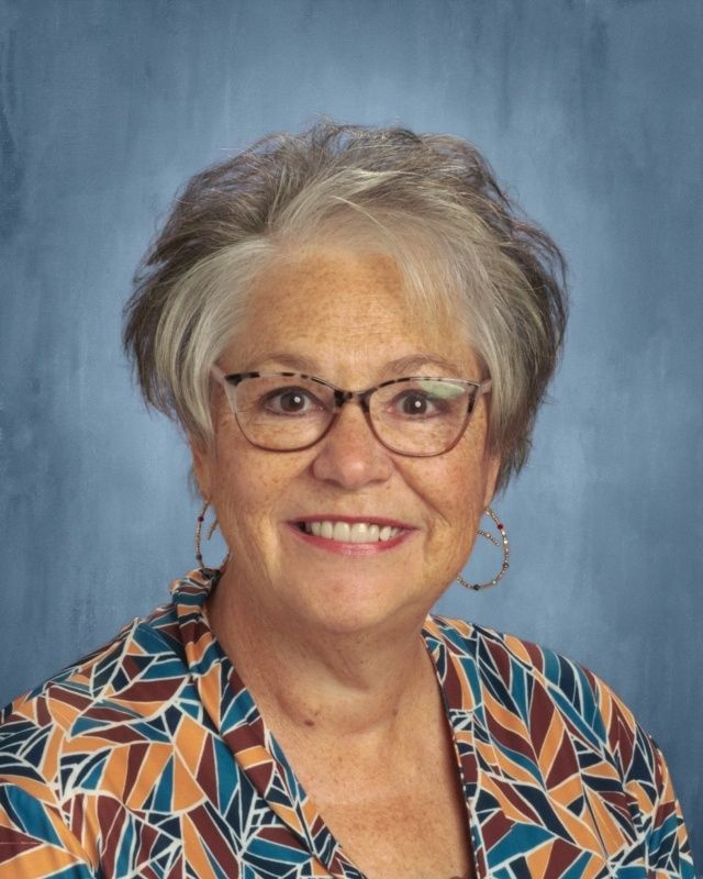 Smiling woman with glasses and graying hair, wearing a patterned top, against a blue backdrop.