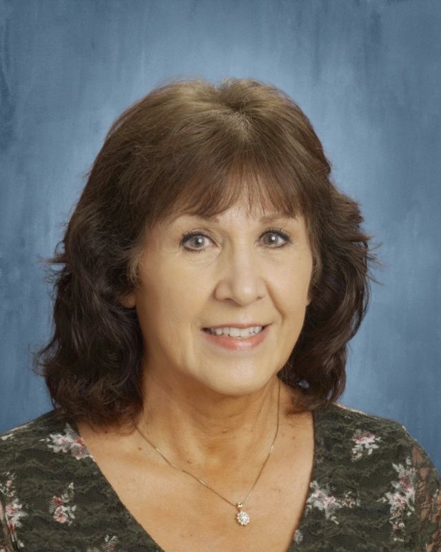 Woman with brown hair, smiling, wearing a floral top and necklace, on a blue background.