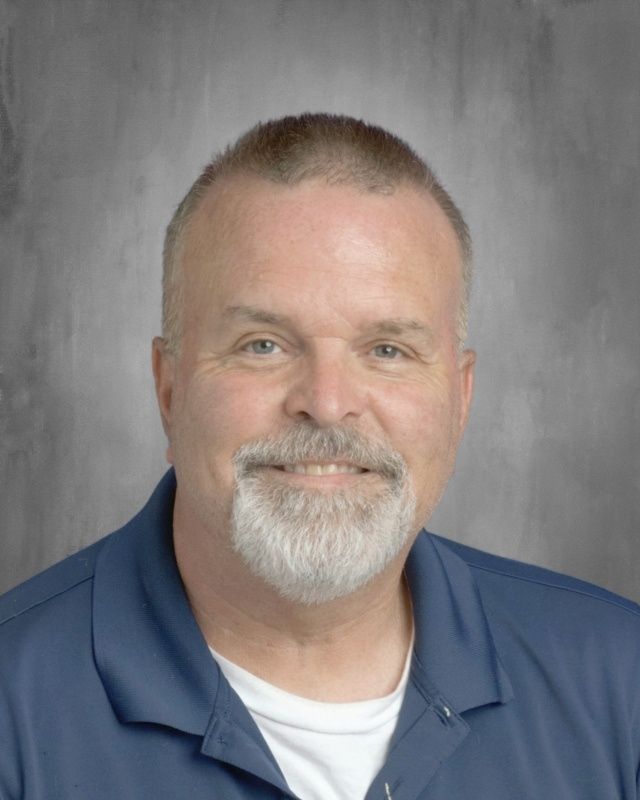 Smiling man with gray beard wearing blue shirt against gray background.