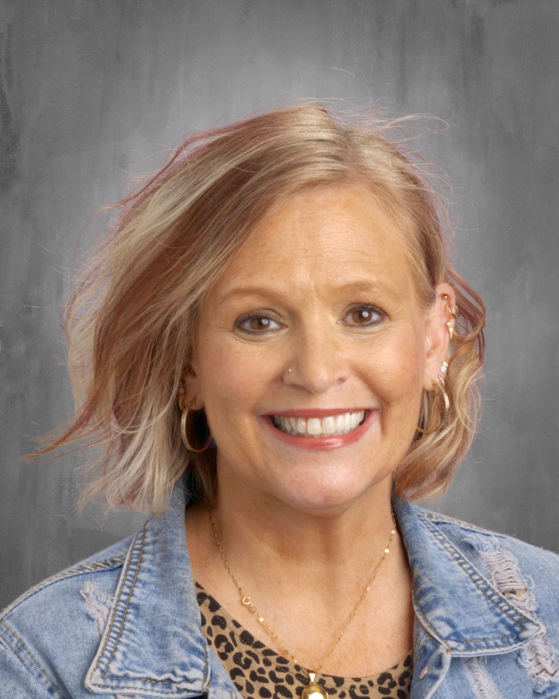Woman with blonde hair, smiling, wearing a denim jacket and leopard print top.