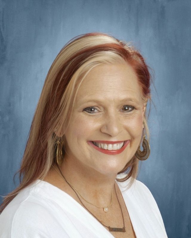 Smiling woman with blonde and red highlights, wearing a white top and jewelry, on a blue background.