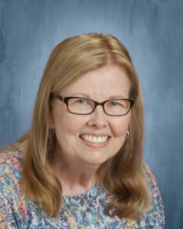 Woman with blonde hair, glasses, smiling, wearing a patterned top; blue background.