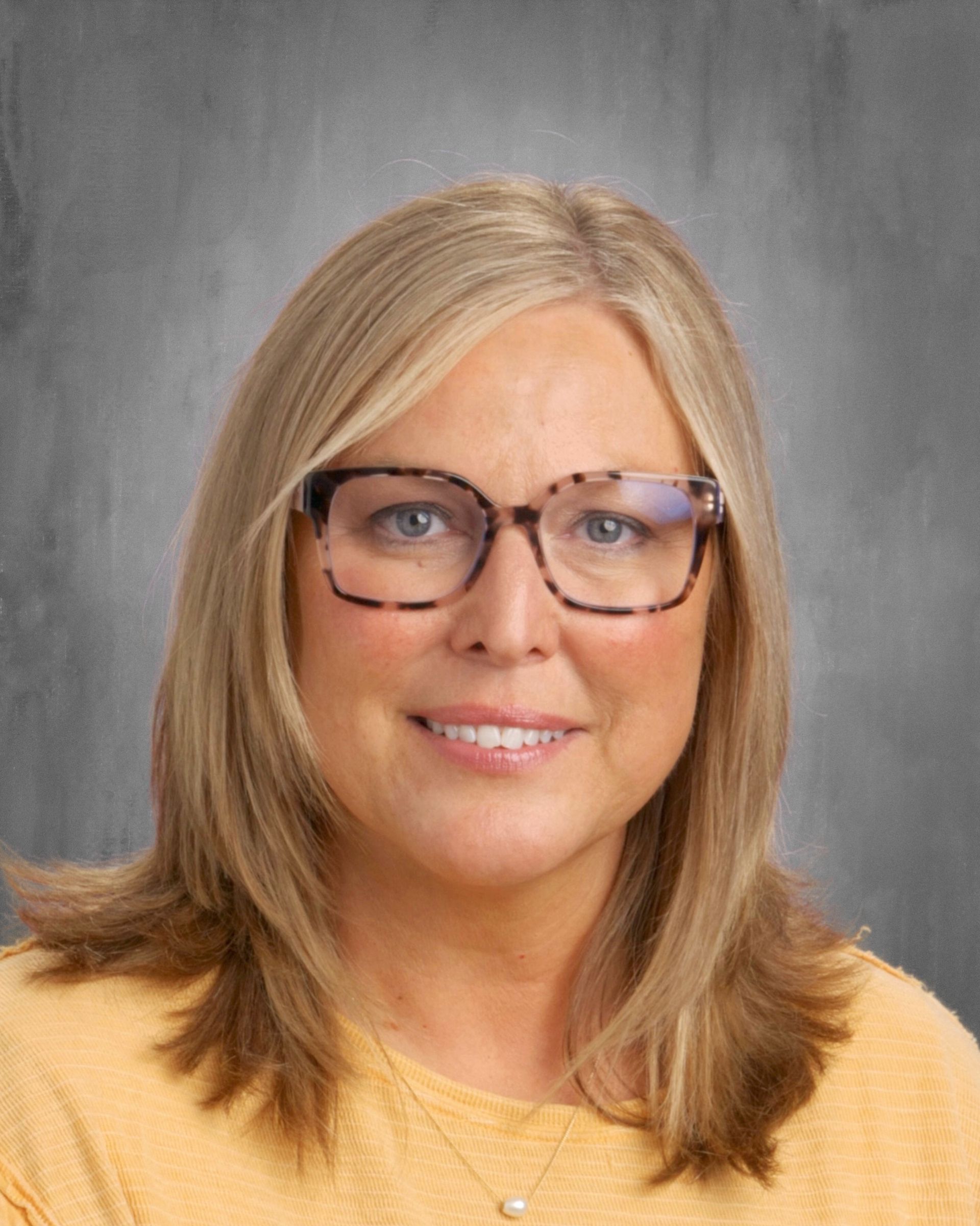 Woman with blonde hair and glasses, wearing a yellow shirt, smiling, studio portrait.