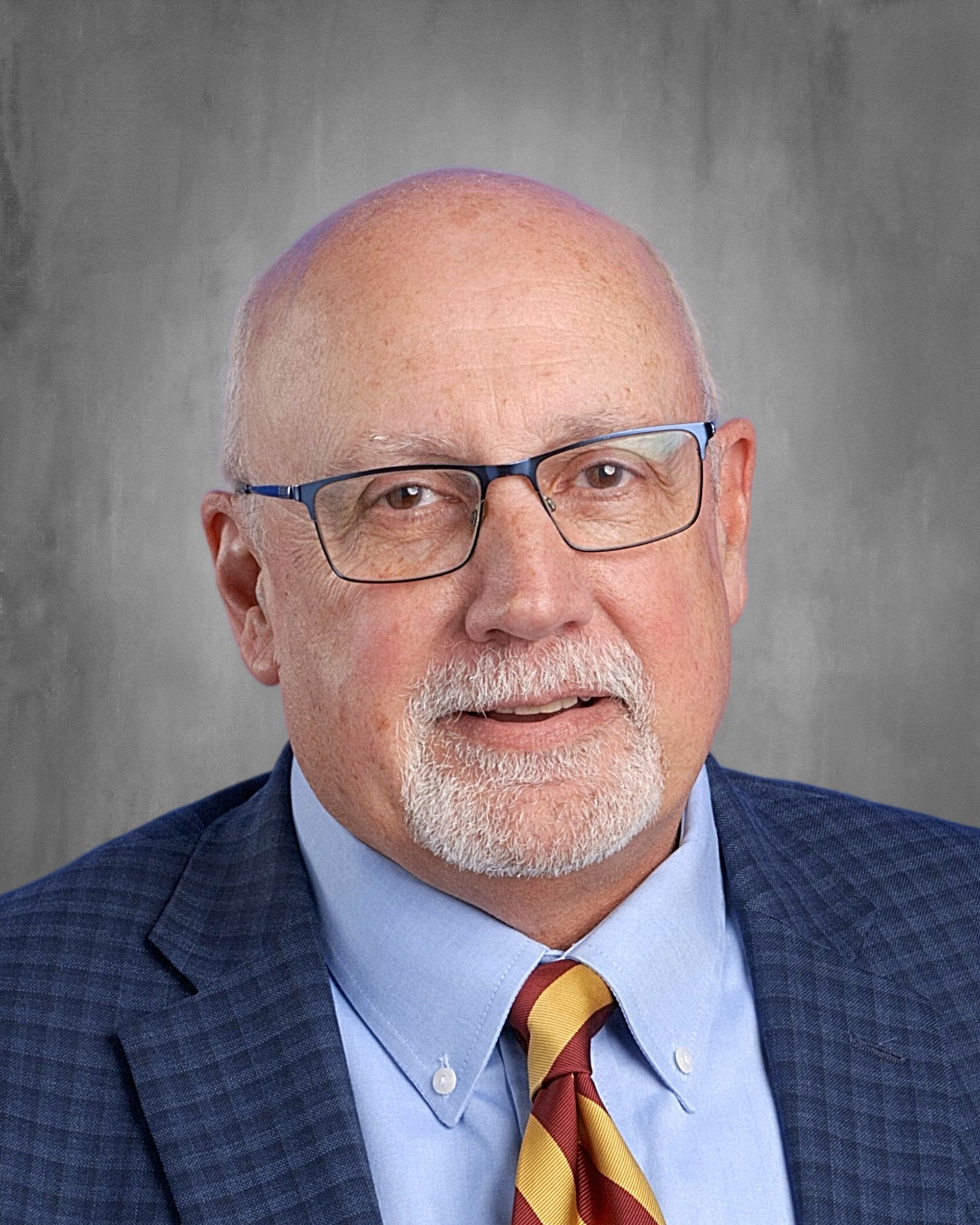 A smiling, older man in glasses and a suit, against a gray background.