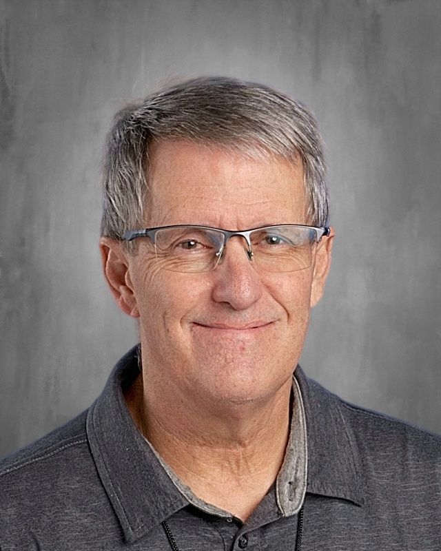 Man with glasses smiling, wearing a blue shirt, against a grey background.