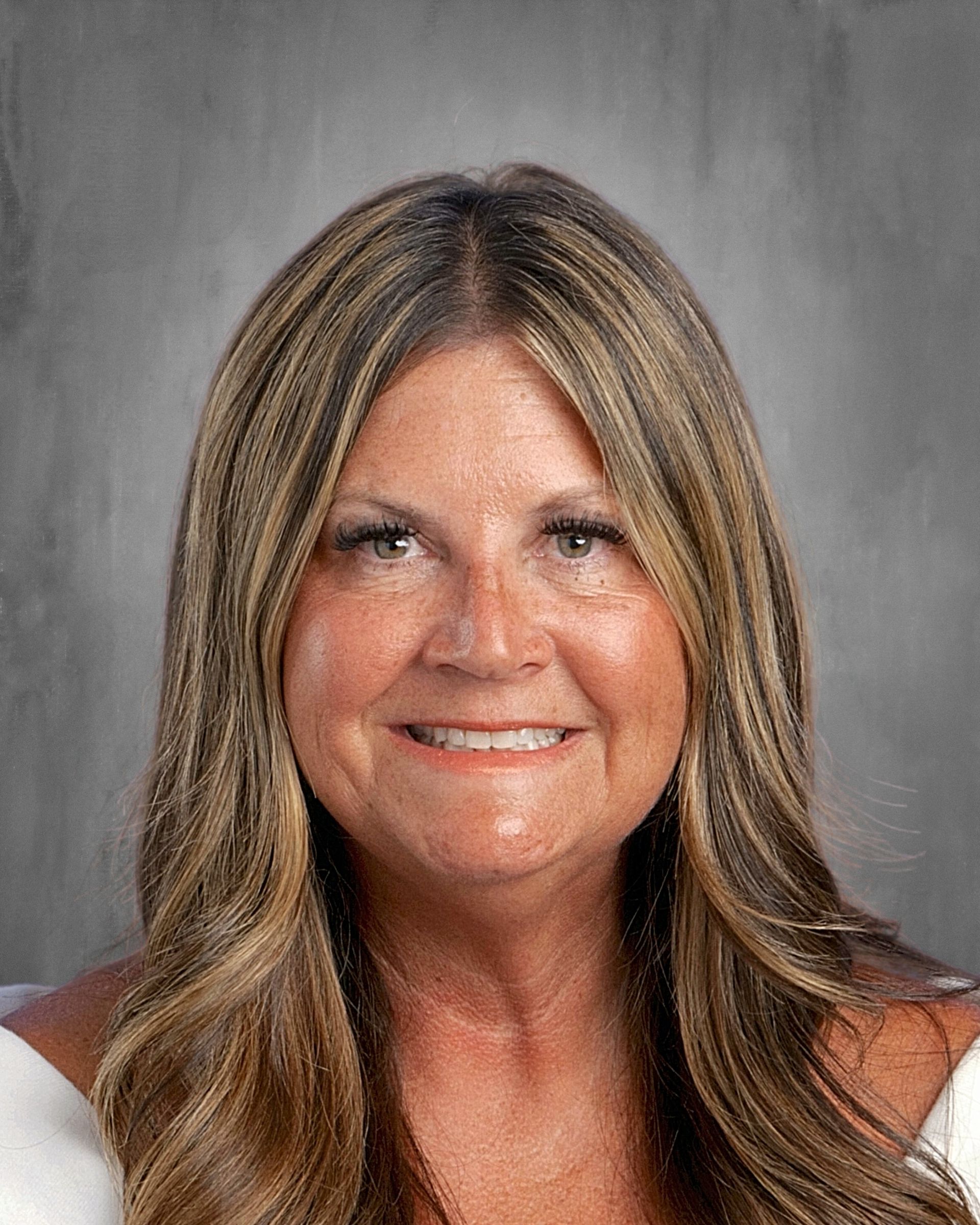 Woman with long, highlighted hair, smiling, wearing a white top against a gray backdrop.