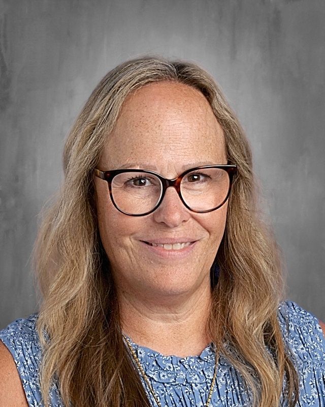 Woman with blonde hair and glasses smiles at the camera. Gray backdrop.