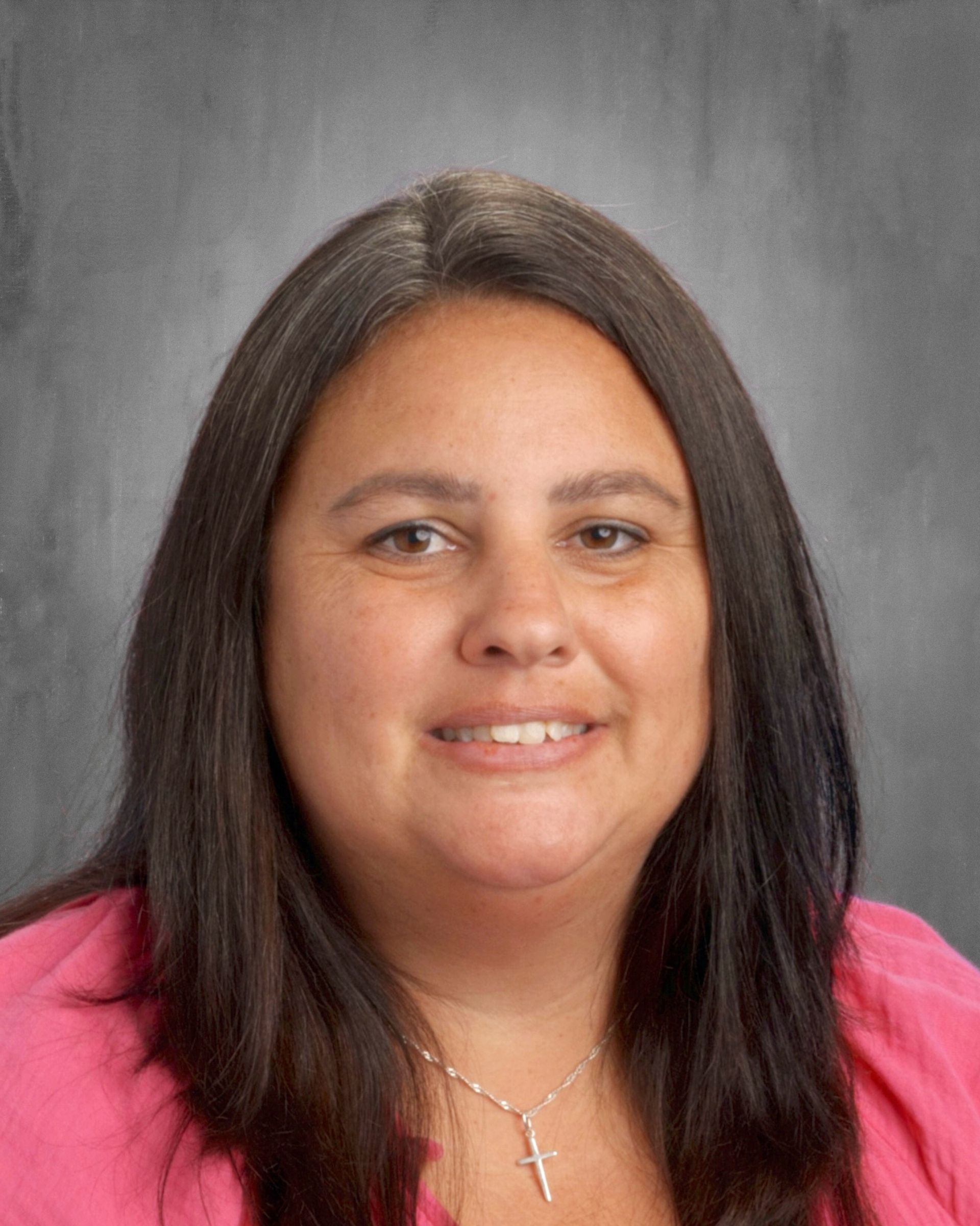 Woman with long dark hair smiles, wearing a pink shirt, against a gray background.