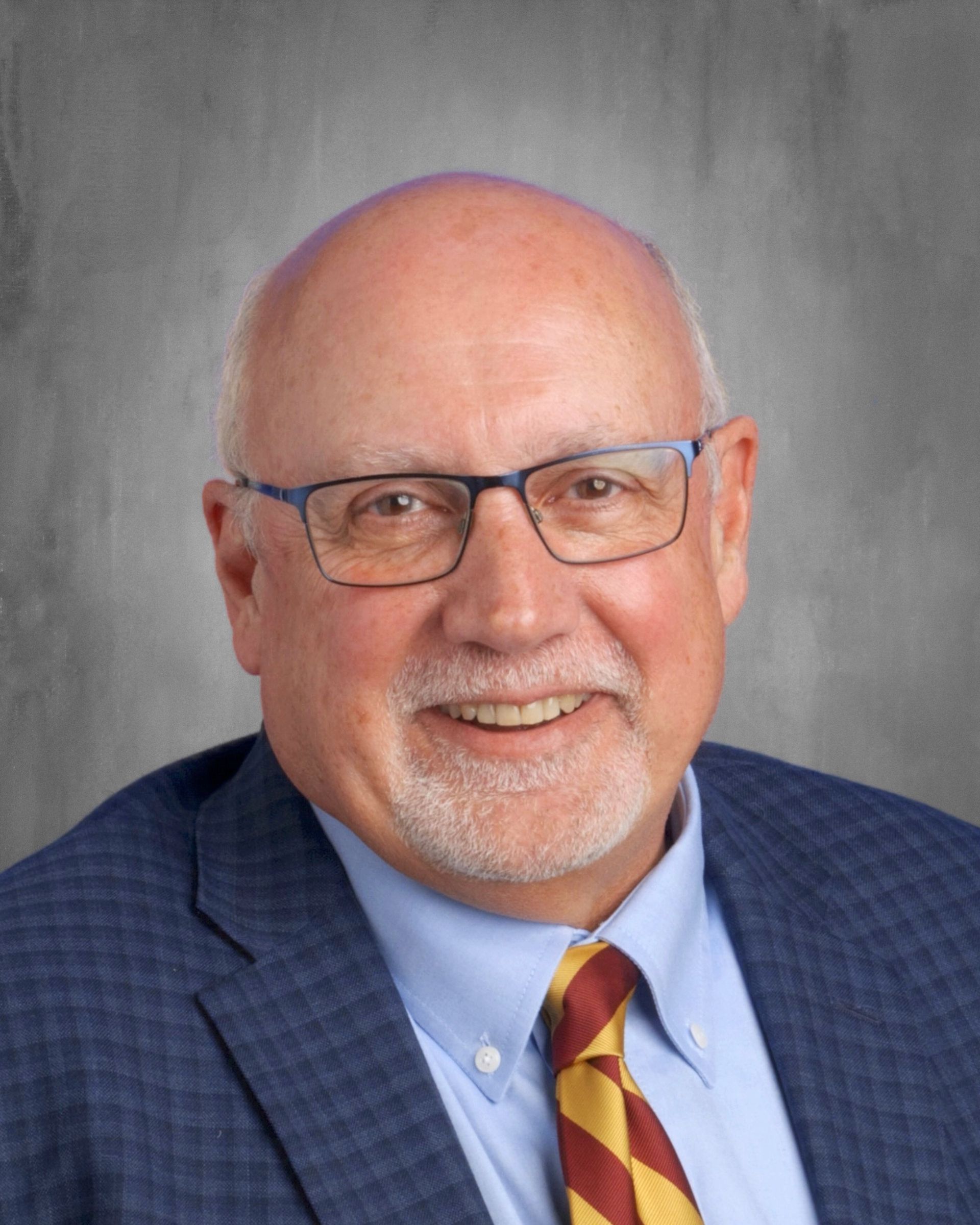Smiling bald man in glasses, blue blazer, colorful tie; against gray background.