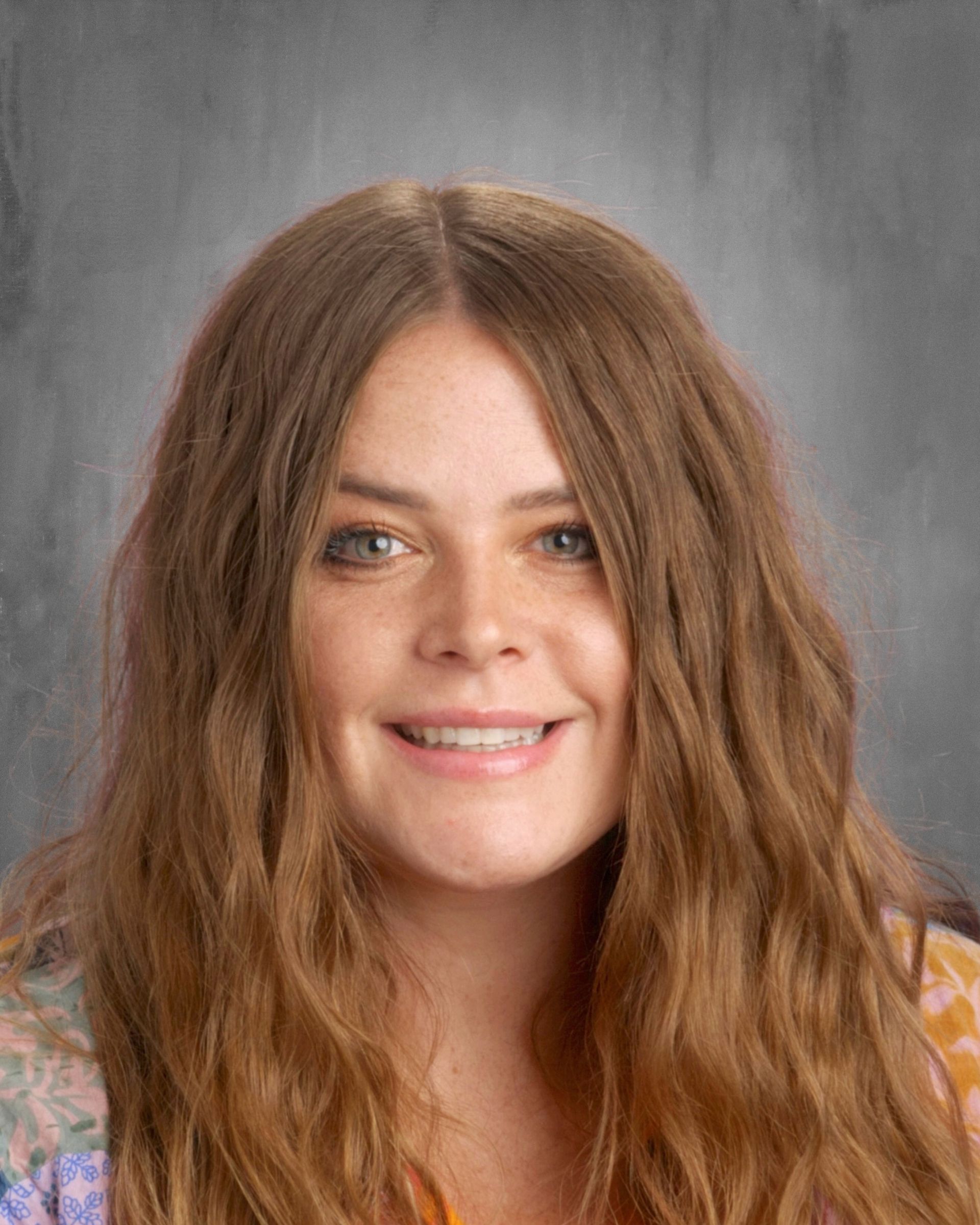 Young woman with long, wavy brown hair smiles at the camera against a gray backdrop.