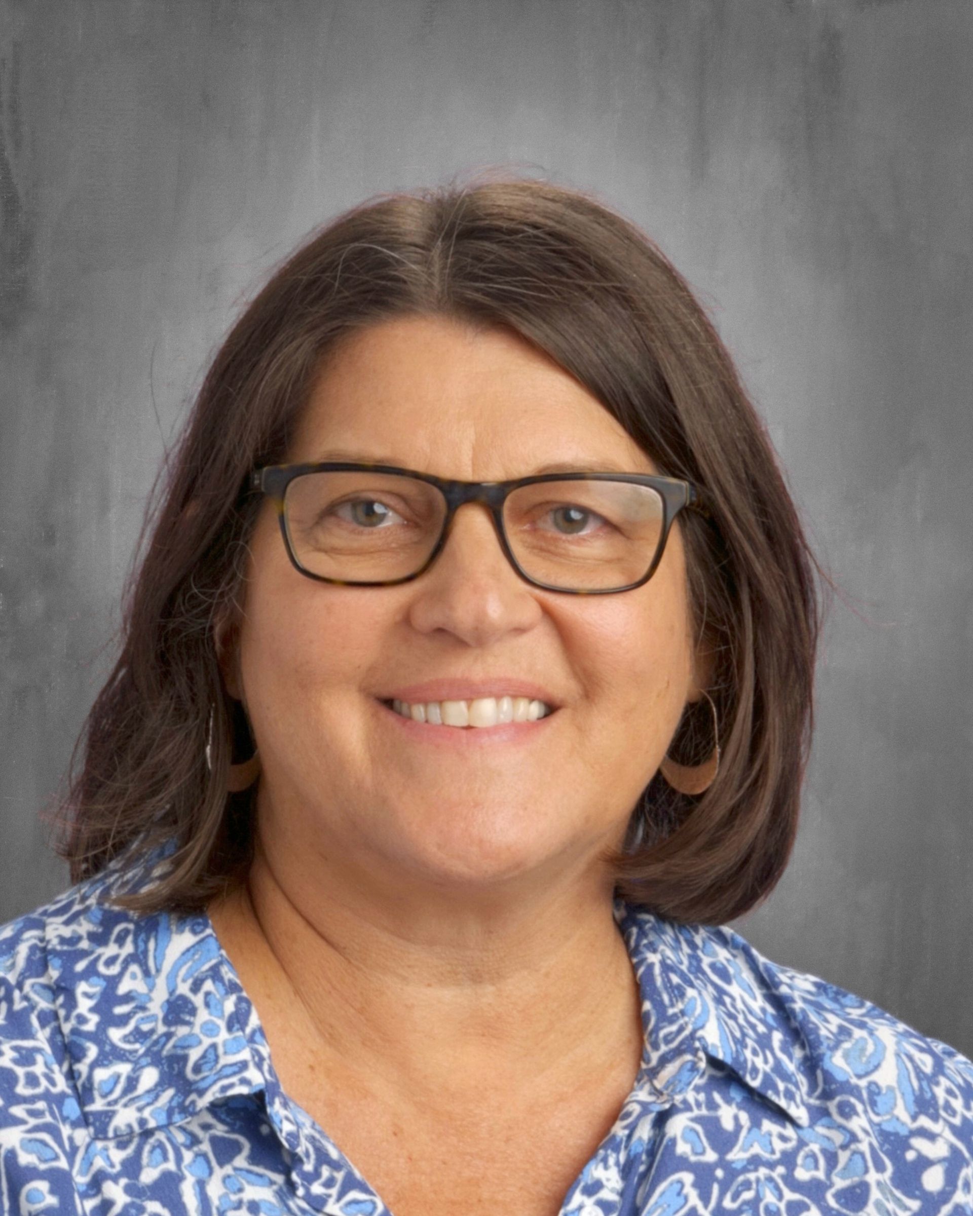 Woman with glasses smiles, wearing a blue floral shirt. Gray background.