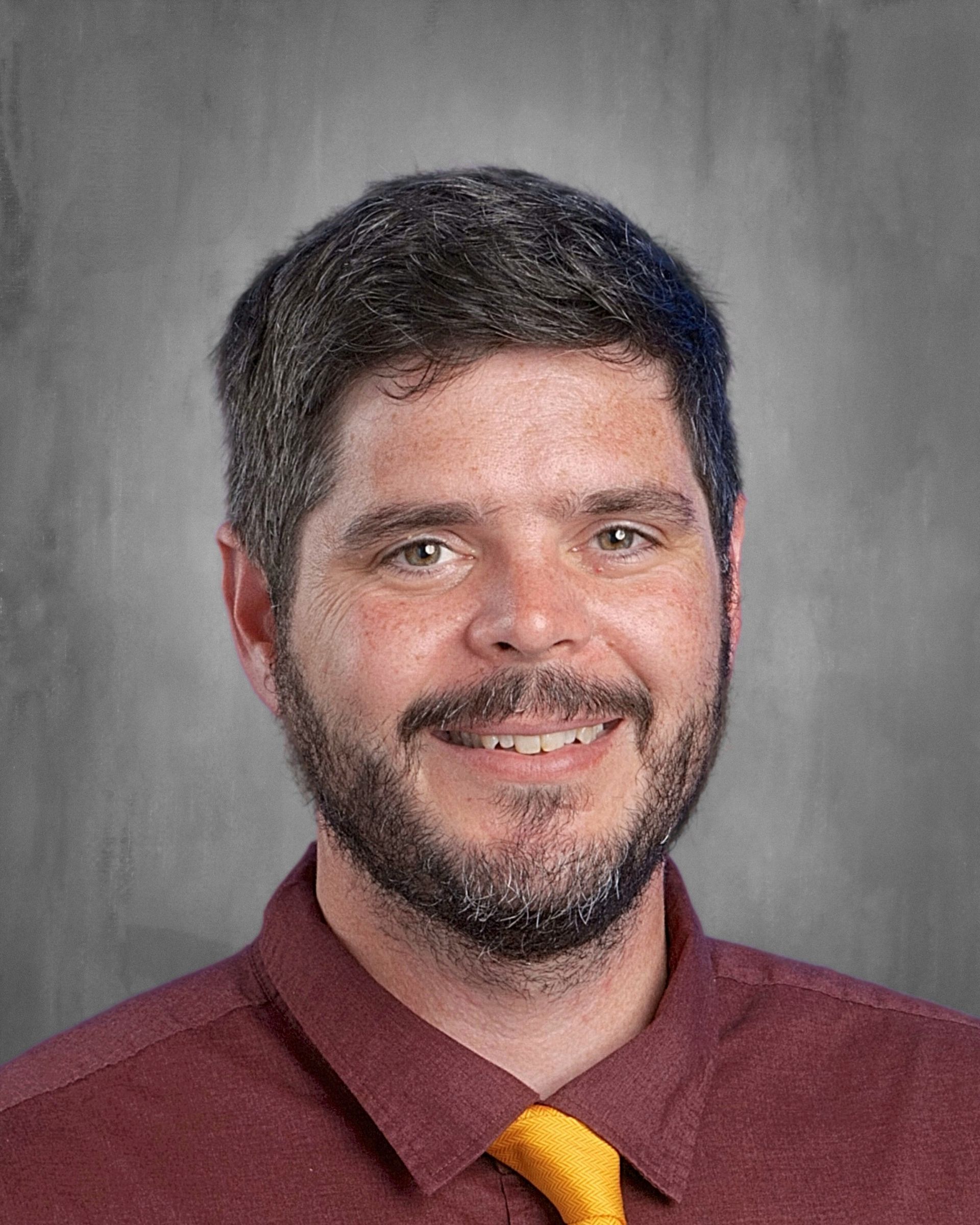 Man with dark hair and beard smiles. He wears a button-up shirt and tie. Gray backdrop.
