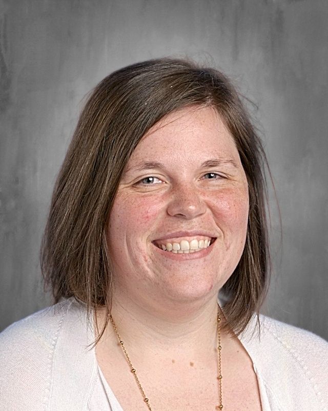 Woman with long brown hair smiles at the camera. She wears a white shirt, against a gray background.