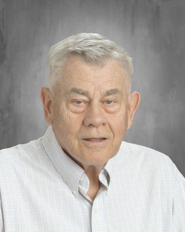 Portrait of an older man with short gray hair, wearing a light blue button-down shirt, neutral background.