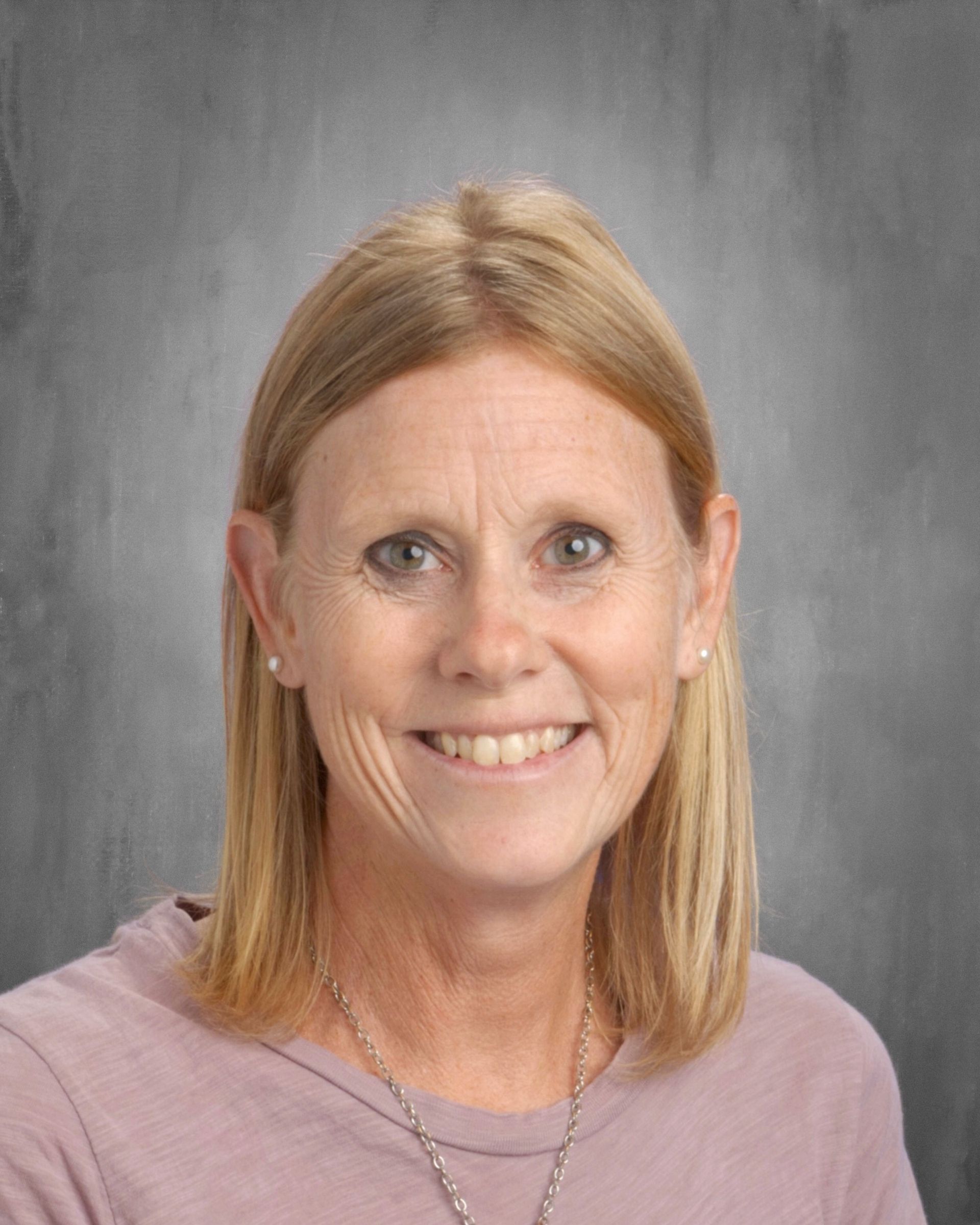 Woman with blonde hair smiles, wearing a light purple shirt, against a gray background.