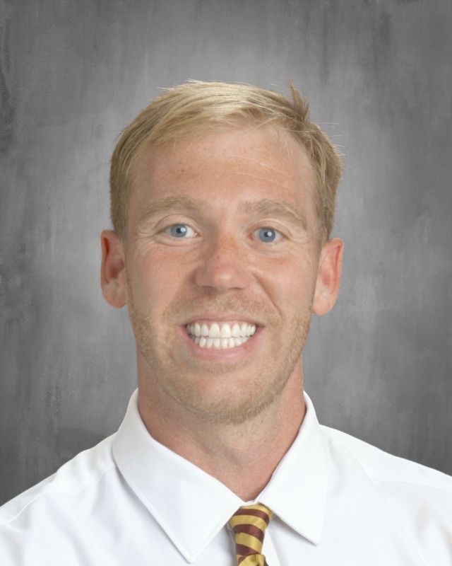 Blond man with a wide smile, wearing a white shirt and tie, against a gray background.