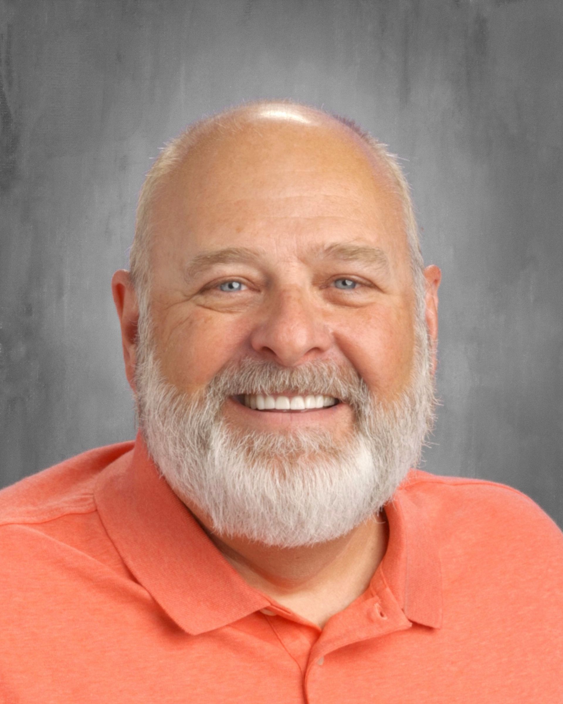 Bald man with white beard smiles, wearing orange polo shirt, grey background.