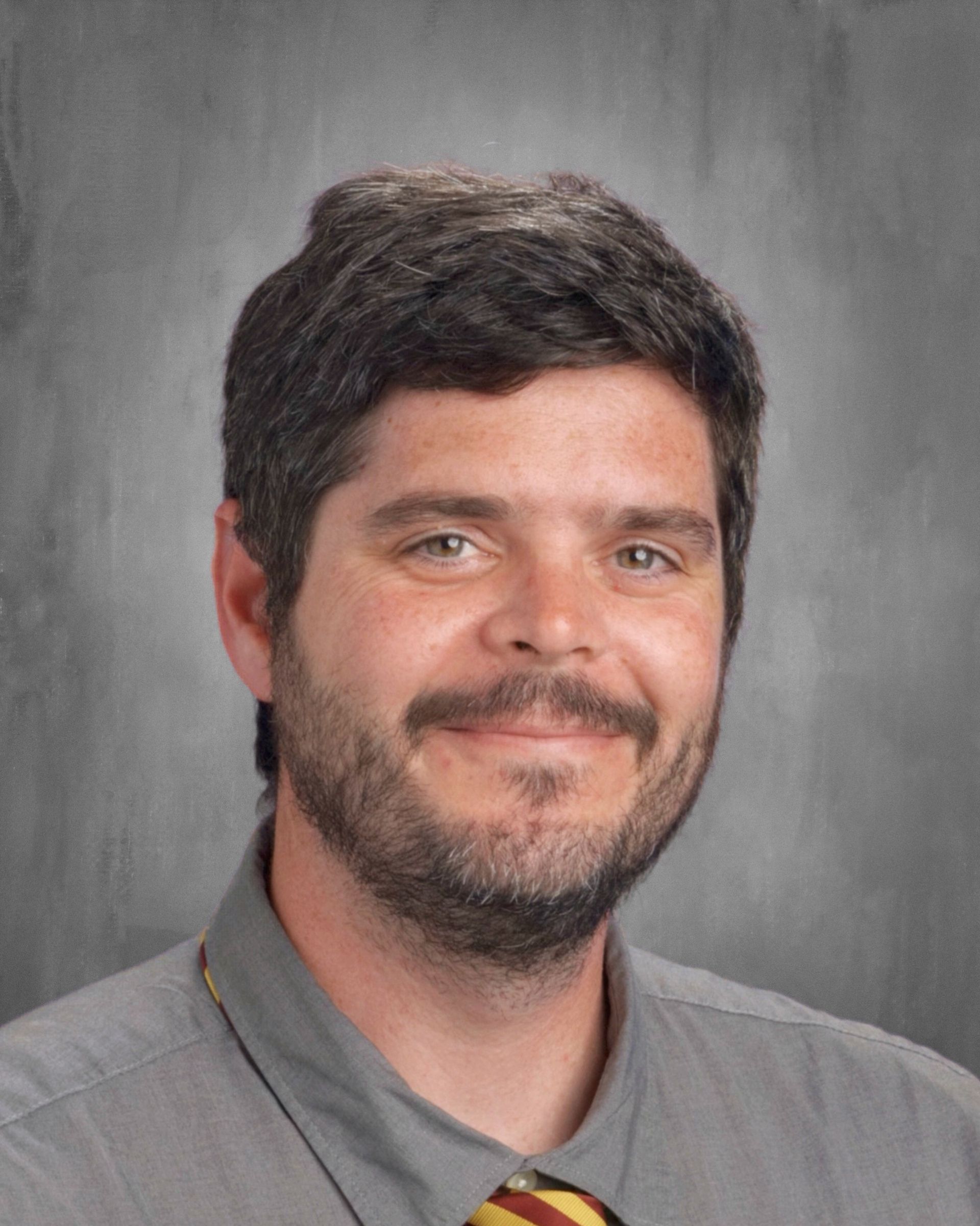 Man with dark hair and beard, wearing a gray shirt and patterned tie, smiling at the camera.