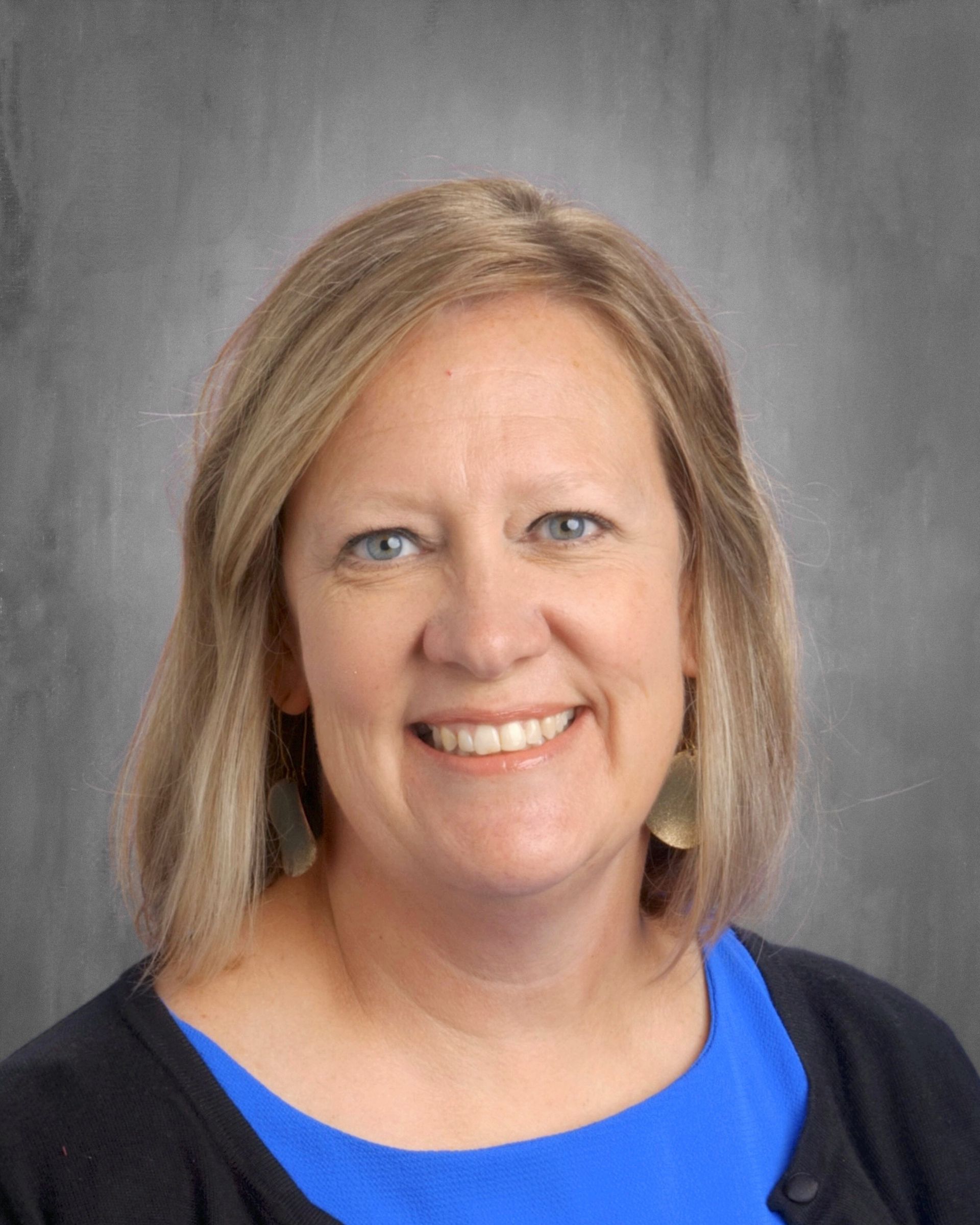 Woman with blonde hair, smiling, blue shirt, black cardigan, gray background.