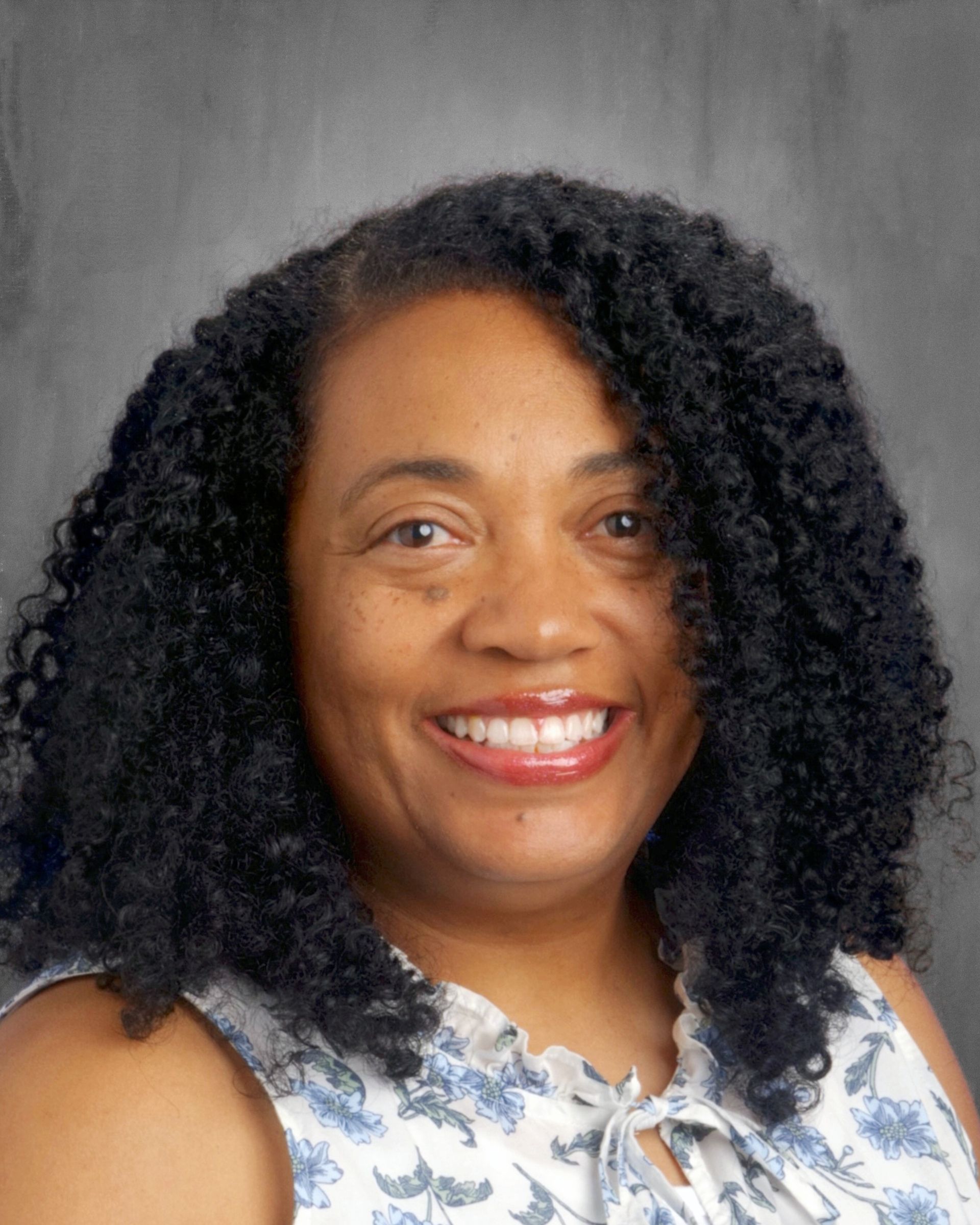 Smiling Black woman with curly black hair, wearing a floral top, against a gray background.