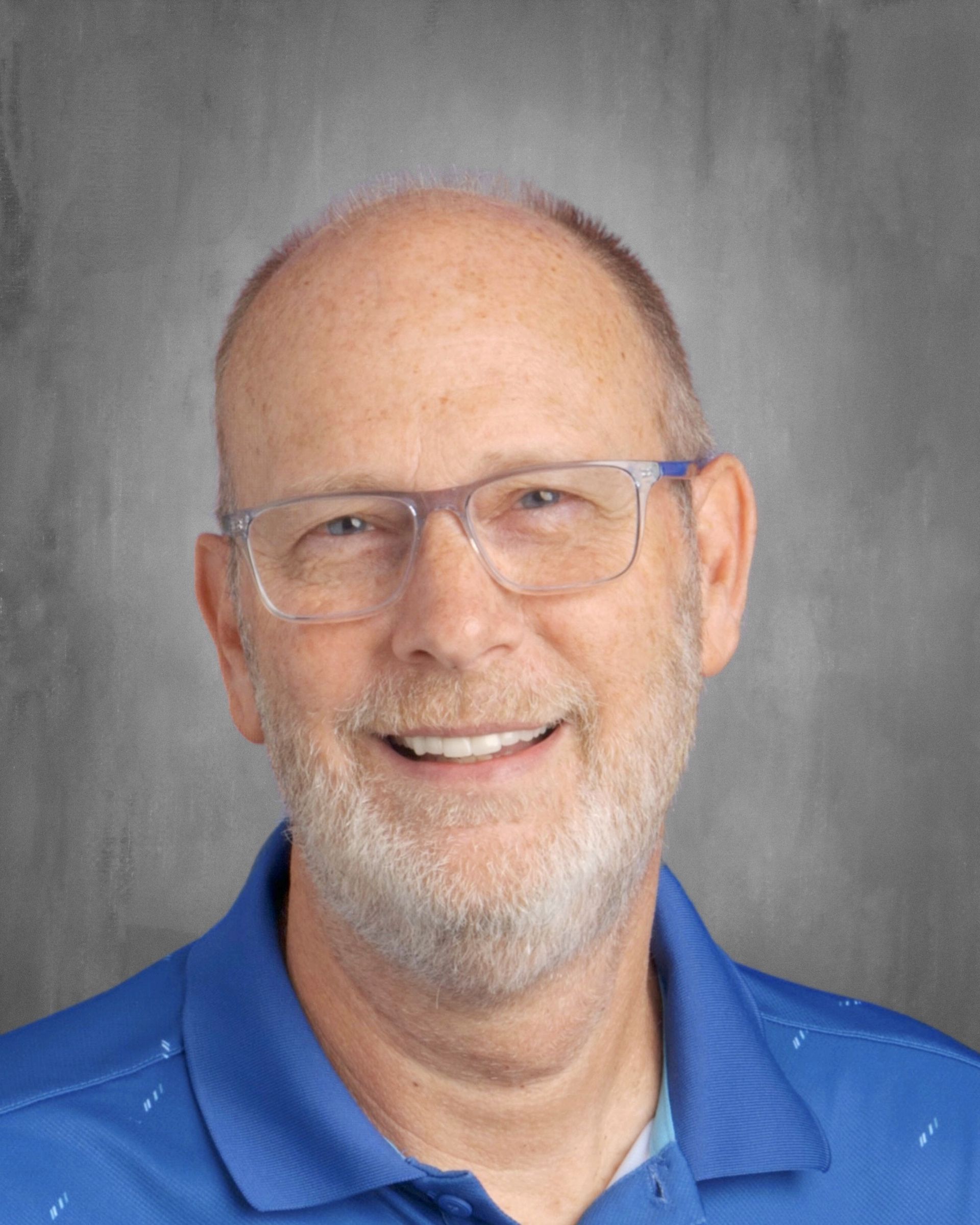 Middle-aged man with glasses, smiling, wearing blue polo shirt, against a gray backdrop.