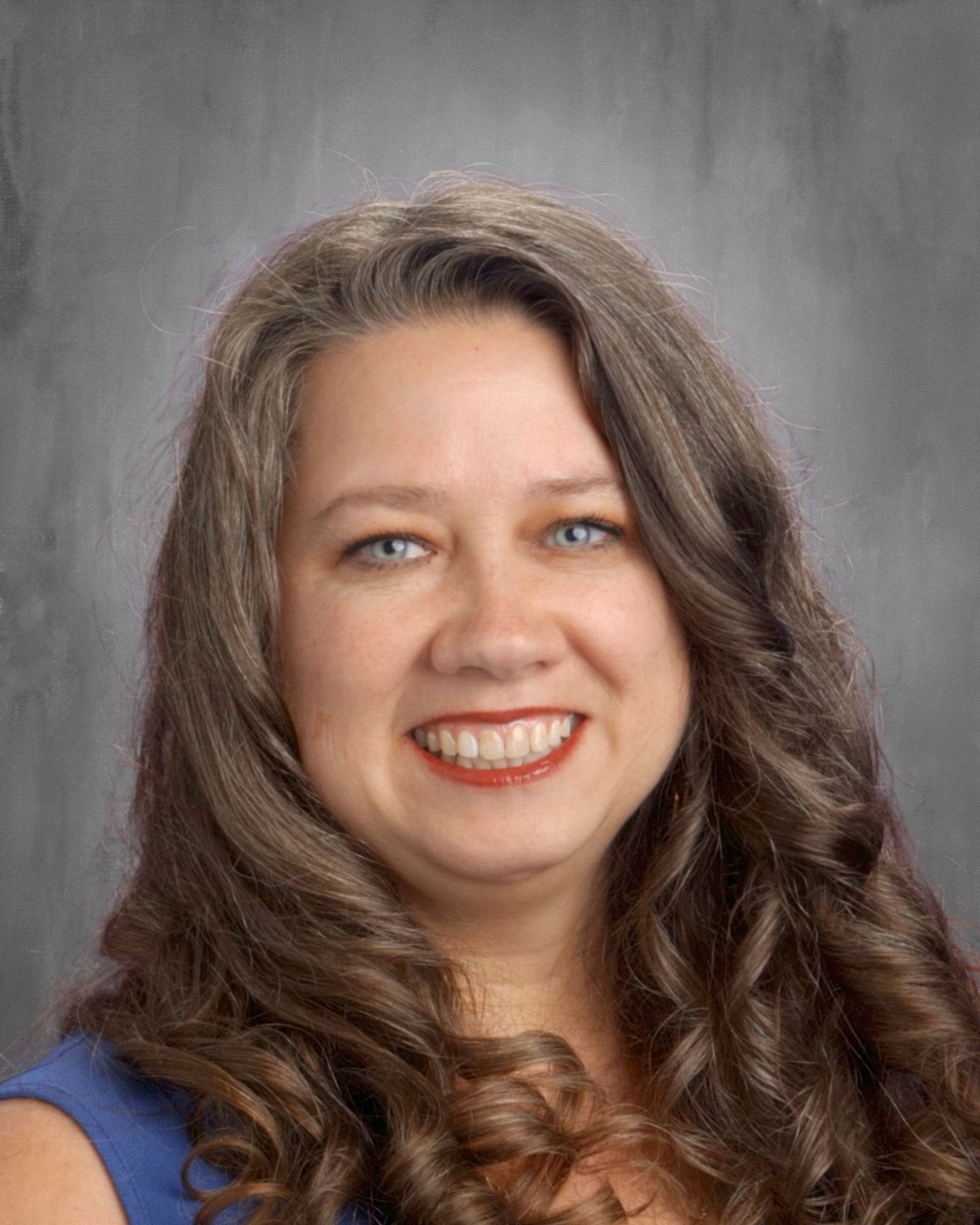 Woman with long wavy brown hair and blue eyes smiles, wearing a blue top, against a gray background.