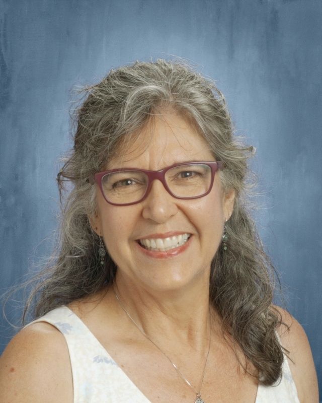 Smiling woman with graying hair, glasses, and a light-colored top, posing in front of a blue background.