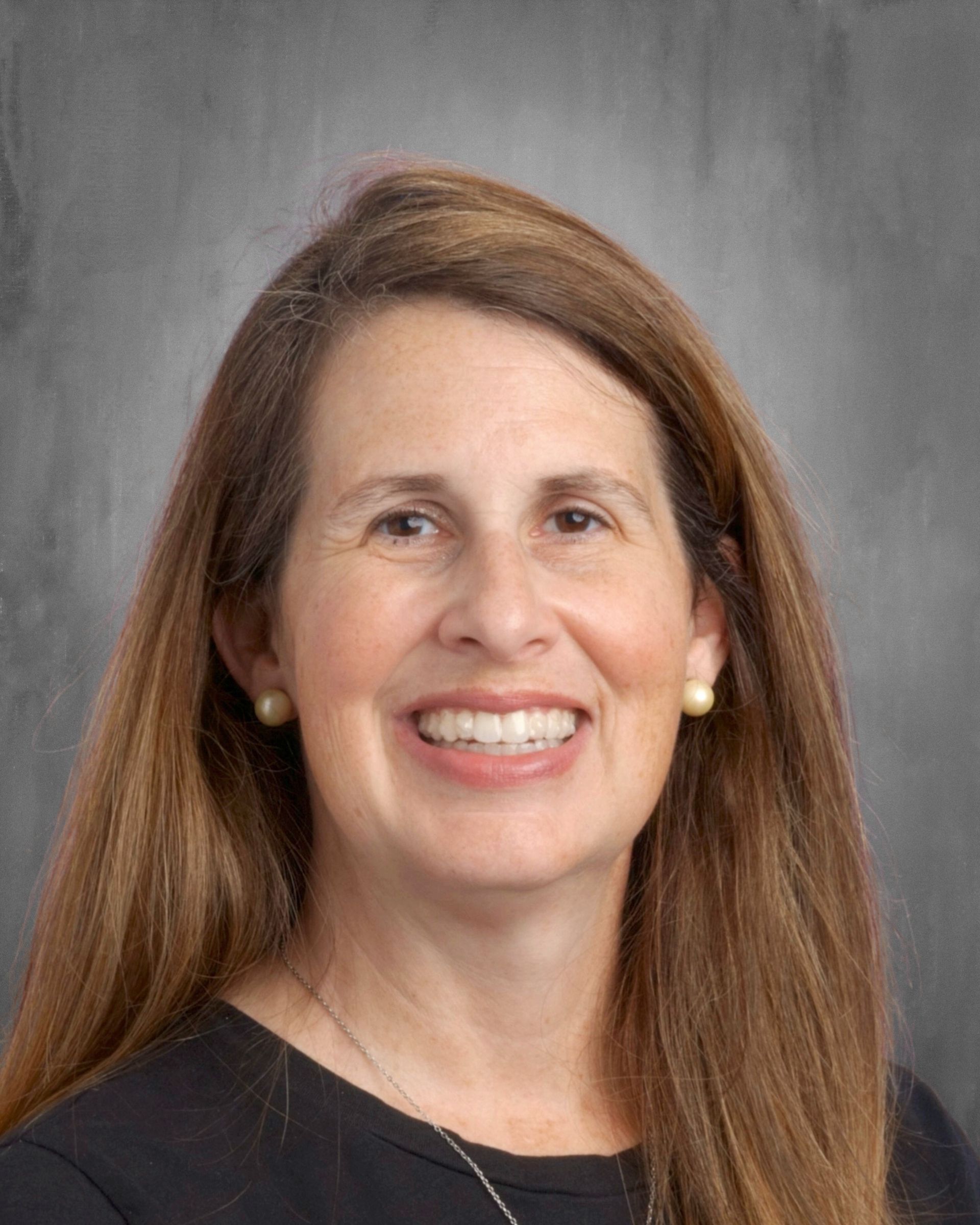 Woman with long brown hair smiling, wearing a black shirt, against a gray background.