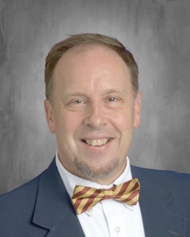 Smiling man in suit, bow tie; gray background.