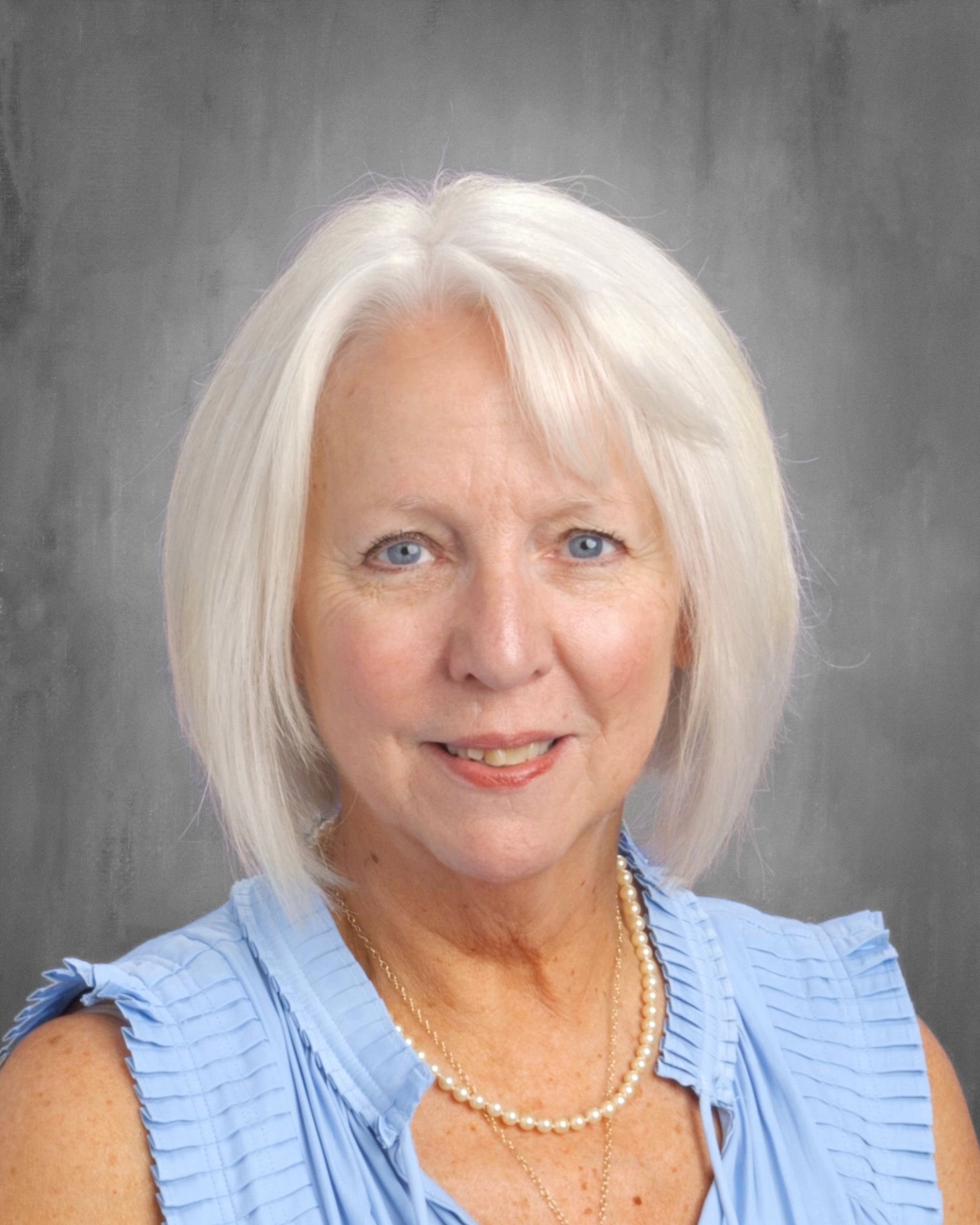 Woman with white hair, smiling, wearing a blue blouse and pearl necklace.