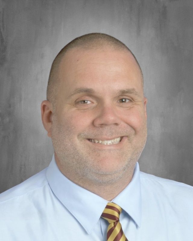 Middle-aged man with shaved head smiles, wearing a blue shirt and maroon/gold striped tie; gray background.