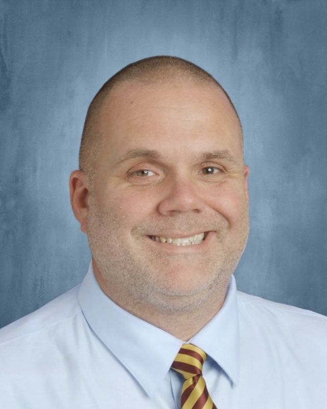 Bald man smiling in a light blue shirt and a maroon and gold striped tie against a blue background.